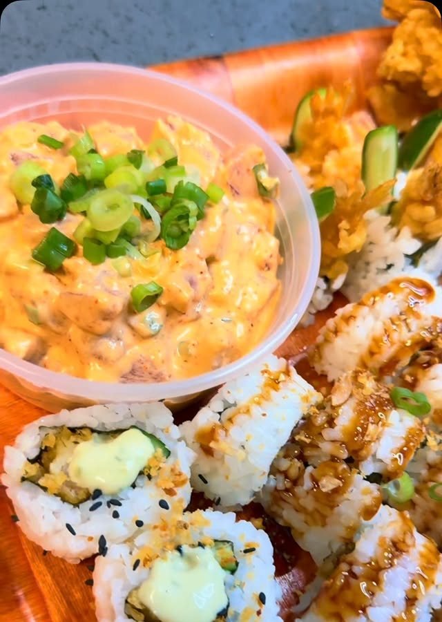 A creamy spicy ahi poke bowl topped with green onions beside crunchy sushi rolls on a wood board